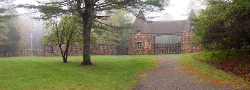 [Several photos stitched together showing a wooden gate stretched between two round, cone-topped stone structures with stone fence and building continuing to the sides.]